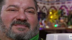 Primer plano de un hombre de mediana edad con barba canosa y expresión amable, bufanda verde y una taza blanca en primer plano inferior, sobre un fondo difuminado con luces y adornos festivos, ¿Quién ganó? Probabilidades entre Eurojackpot y Cupón Extra