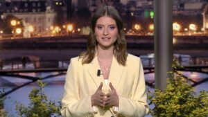 Presentadora en un estudio de televisión con blazer amarillo pálido y collares, micrófono de solapa visible y manos gesticulando frente a un fondo urbano nocturno con puente y luces reflejadas y plantas en primer plano, ¿Quién te hará millonario: El Gordo o Euro Dreams?