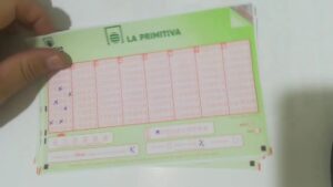 Formulario verde de La Primitiva sobre una superficie blanca sostenido por una mano, con varias casillas marcadas con cruces a bolígrafo y campos de datos cumplimentados; Descubre quién ganó: ¿La Primitiva o el Cuponazo tiene más suerte?